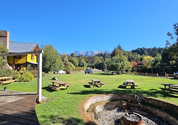 Un jardín con mesas de picnic de madera, un fogón de ladrillo en primer plano, una cabaña amarilla y montañas bajo un cielo despejado