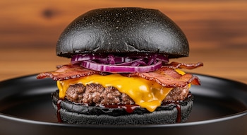 Hamburguesa con pan brioche negro, carne, queso cheddar amarillo fundido, tiras de panceta crujiente y rodajas de cebolla morada en un plato oscuro con fondo de madera.
