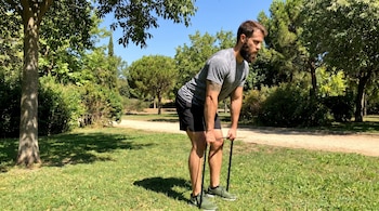 Hombre barbudo inclinado, sostiene banda elástica negra. Viste camiseta gris, pantalón corto negro, zapatillas verdes. En parque con césped y árboles.