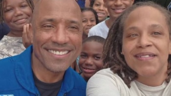 Primer plano de un grupo diverso de personas sonriendo; un hombre calvo con camisa azul a la izquierda, una mujer con rastas a la derecha, y varios niños y jóvenes detrás