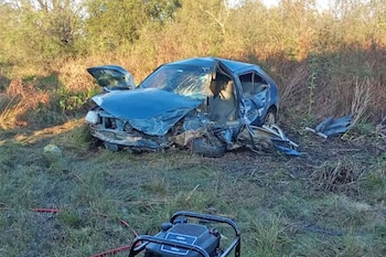 Un vehículo azul con el frente destruido y la puerta abierta yace sobre el pasto junto a un generador portátil