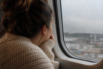 Plano en detalle de una mujer con suéter beige mirando por la ventanilla de un avión cubierta por gotas de lluvia.