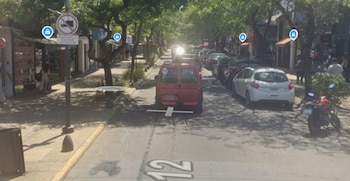 Una calle arbolada con un vehículo rojo en circulación, automóviles estacionados, locales comerciales y el número doce en la calzada
