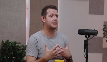 Un hombre con camiseta gris habla frente a un micrófono negro en un estudio con paneles acústicos, una planta verde y una taza amarilla
