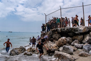 Un grupo de hombres en una orilla rocosa, algunos en el mar, otros trepando una valla metálica con alambre de púas. Varias barcas en el horizonte