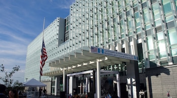 Fachada de un edificio moderno de cristal y metal con la bandera de Estados Unidos a media asta, un cartel sobre la entrada y un evento exterior con carpas y escenario.