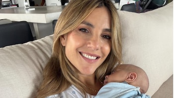 Primer plano de María Belén Ludueña sonriendo a cámara, con cabello castaño claro, sosteniendo a su bebé Vito dormido envuelto en una manta azul