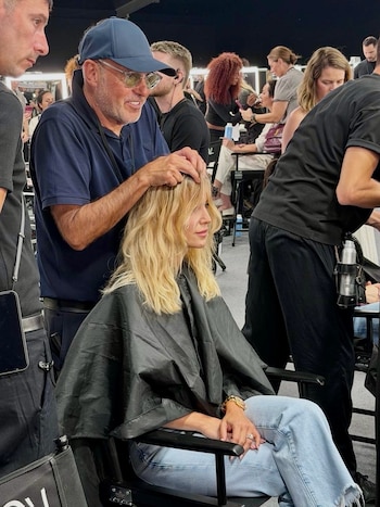 Luisana Lopilato sentada con una capa negra mientras un estilista con gorra le acomoda el cabello rubio en un salón lleno de gente