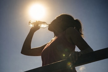 Silueta de una mujer bebiendo agua de una botella de plástico. El sol brilla en el cielo azul. Gotas de agua caen. Una barandilla oscura en primer plano.
