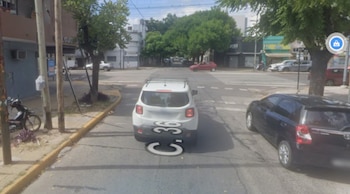 Vista aérea de una intersección urbana con varios autos estacionados y en movimiento, árboles y edificios de baja altura. Un auto blanco tiene un marcador de Google Street View