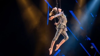 Persona con traje brillante plateado y pedrería suspendida por dos cintas negras, con luces de escenario azules y amarillas de fondo