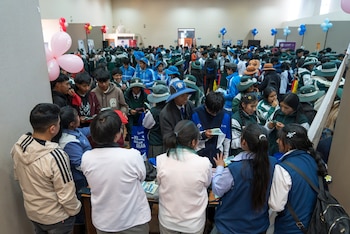 Decenas de jóvenes con uniformes escolares y sombreros se reúnen alrededor de mesas con folletos en un salón amplio decorado con globos