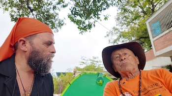 Primer plano de dos hombres, uno con gorro naranja y barba, el otro con sombrero marrón y camiseta naranja. Fondo con tienda de campaña verde, árboles y un edificio.