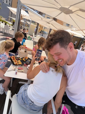 Cinco personas, incluyendo Michael Bublé y Luisana Lopilato, en una mesa exterior con sombrillas. Hay perritos calientes, papas fritas y un teléfono