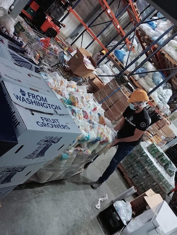 Hombre con casco y mascarilla mueve una paleta de cajas y bolsas en un almacén. Cajas "FROM WASHINGTON FRUIT GROWERS". Estanterías y mercancía al fondo