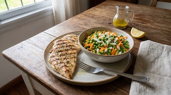 Un plato sobre una mesa de madera contiene pechugas de pollo grillé y un tazón de ensalada de arvejas, zanahoria y cebolla, con un limón y aderezo.