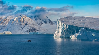 Un barco pequeño con personas navega por aguas azules cerca de grandes icebergs y acantilados de hielo, con montañas nevadas y cielo azul de fondo.