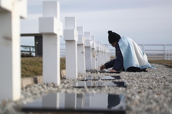 Una persona inclinada junto a una hilera de cruces blancas en un cementerio, cubierta con una manta con los colores de la bandera argentina
