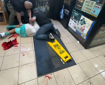 Vista aérea de un hombre en el suelo, inmovilizado por dos personas, con un charco de sangre roja brillante sobre baldosas claras y una señal de "Piso mojado"