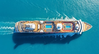 Vista aérea de un crucero blanco y beige con piscinas azules y tumbonas en sus cubiertas superiores, navegando en aguas turquesas brillantes dejando una estela blanca.
