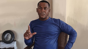 Ilustración de un hombre con camiseta azul sentado en una silla, con la mano derecha alzada junto a una fotografía enmarcada.