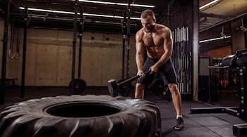 Un hombre sin camiseta, musculoso y barbudo, golpea un neumático grande con un martillo en un gimnasio de CrossFit. El hombre está sudoroso.