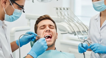 Un hombre con boca abierta en silla dental siendo examinado por un dentista con mascarilla y guantes, asistido por otra persona.