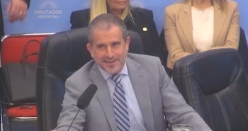 Un hombre con traje gris y corbata azul a rayas sonríe sentado frente a un micrófono ante una pared con la frase Diputados Argentina