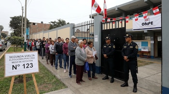 Larga fila de personas espera votar en una mesa electoral en Perú. Dos policías custodian la entrada. Se ven banderas peruanas y un cartel de la ONPE.