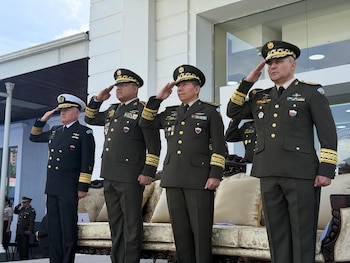 Cuatro militares de pie en uniforme, uno de la marina y tres del ejército, saludan. Llevan gorras con insignias doradas. Al fondo, un edificio blanco