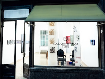 Interior de sala luminosa con paredes blancas y suelo de baldosas claras, mesa de madera, sillas modernas, cuadros, tapices coloridos y dos sillas negras ornamentadas