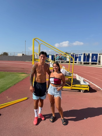 Hombre y mujer posan de pie en una pista de atletismo. Detrás hay gradas amarillas, vallas metálicas y edificios modulares bajo un cielo azul despejado