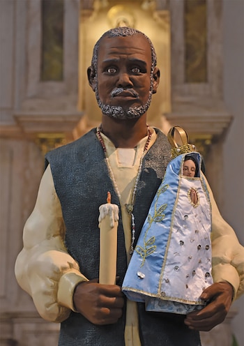 Estatua de un hombre con piel oscura y barba gris, viste camisa clara y chaleco oscuro, sostiene una vela y una pequeña virgen vestida de azul y blanco