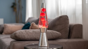 Una lámpara de lava con cera roja brillante y líquido transparente, con base y tapa plateadas, colocada sobre una mesa redonda frente a un sofá gris.