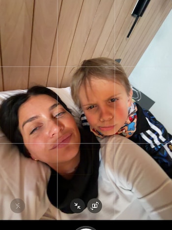 Una mujer de cabello oscuro y un niño rubio acostados en una cama con almohadas blancas. Detrás se ve una pared de madera. Controles de cámara