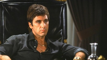 Al Pacino, en primer plano medio, con camisa oscura y cadena de oro, sentado en una silla de cuero negro con detalles dorados, con una expresión seria