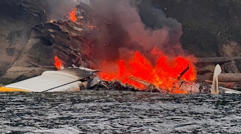 Restos de una aeronave, con fuego y humo, parcialmente sumergidos en el agua cerca de una costa rocosa con vegetación.