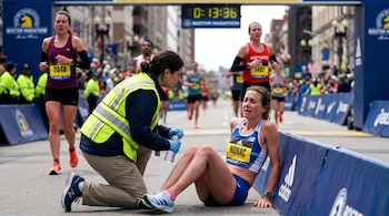 Una corredora sentada en el suelo junto a una barrera recibe asistencia de personal voluntario tras cruzar la meta de un maratón.
