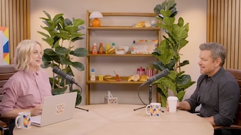 Amy Poehler y Matt Damon en una mesa con micrófonos, un portátil, tazas y una taza de café. Detrás, un estante con decoraciones de comida y plantas