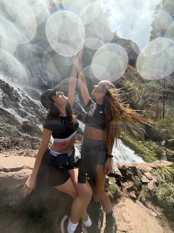 Dos mujeres sonríen y extienden sus brazos hacia arriba, tocándose las manos, frente a una cascada natural. Llevan ropa deportiva oscura