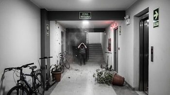 Pasillo de edificio con escombros y humo. Un hombre cubre su cabeza. Hay dos bicicletas, dos scooters, macetas, señales de seguridad y salida, escaleras y ascensor