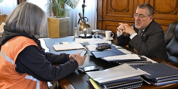 Dos personas, una con chaleco naranja y la otra con traje, sentadas en un escritorio con documentos, teléfonos móviles, una lámpara y un reloj de arena