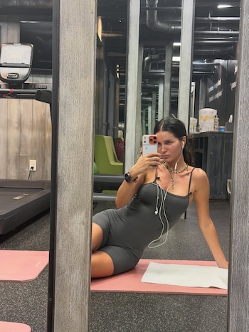 Zaira Nara, sentada en una colchoneta rosa, se toma una selfie en el espejo de un gimnasio, vestida con un enterito gris y auriculares blancos