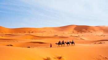 Cuatro personas montan camellos, una persona los guía a pie. Un desierto con dunas de arena naranja y cielo azul claro se extiende al fondo