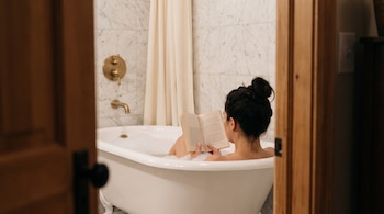 Mujer de espaldas con moño leyendo un libro en bañera blanca con patas y espuma, rodeada de paredes de mármol, grifos de latón y cortina beige.