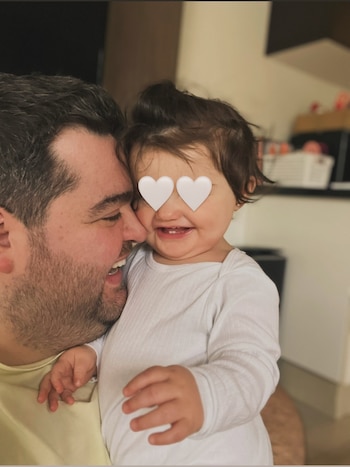 Hombre barbudo con camiseta clara abraza a bebé de cabello oscuro con camiseta blanca. Ambos sonríen. Ojos de la bebé con corazones blancos