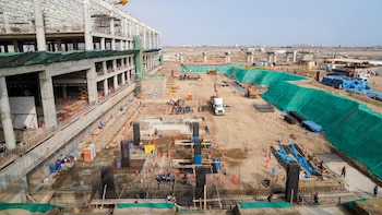 Un sitio de construcción extenso con un edificio en obra, andamios, grúas, camiones, excavadoras, trabajadores, barras de acero, conos y tierra