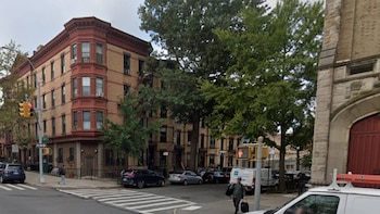 Vista de una calle urbana con un edificio de ladrillo y ventanales en esquina, un paso de cebra, coches, furgonetas, árboles y peatones