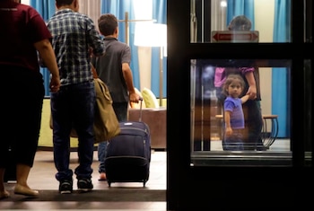 Una niña pequeña de camiseta morada se ve a través de un cristal, tomada de la mano con un adulto; al fondo, tres personas con equipaje se alejan