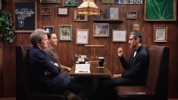 Sundar Pichai, con gafas y barba, gesticula mientras conversa con dos hombres sentados frente a él en una mesa de madera de un pub. Hay vasos de cerveza y cuadros decorativos